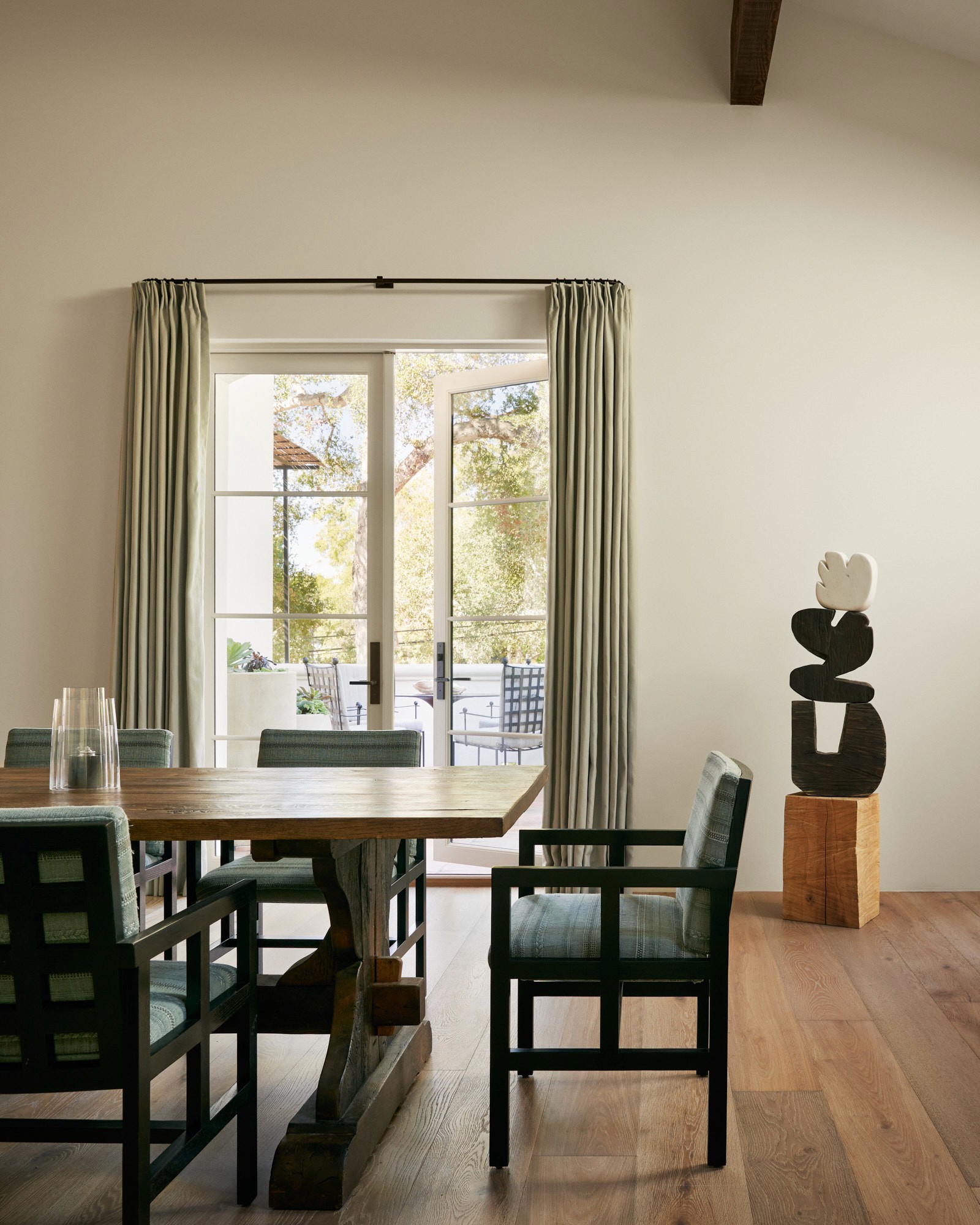 A dining area with a weathered trestle table and dark-framed armchairs upholstered in green. An abstract wood sculpture on a timber pedestal stands in the corner. French doors with sage curtains open to the terrace beyond.