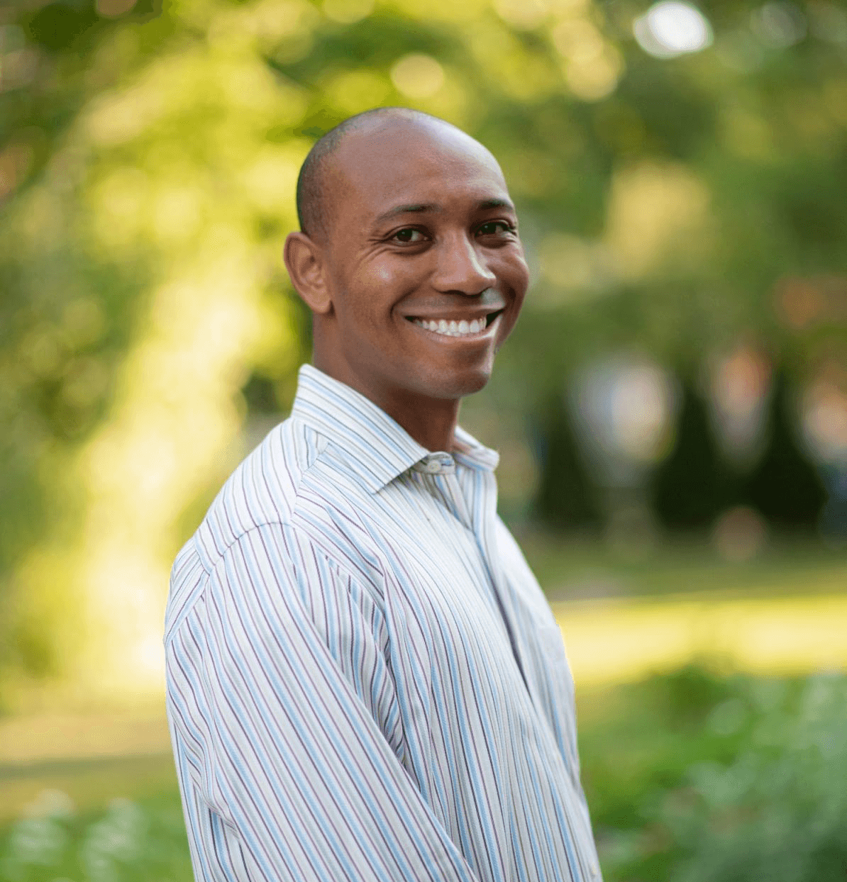 A man smiles while standing outdoors in a park, surrounded by greenery and sunlight.