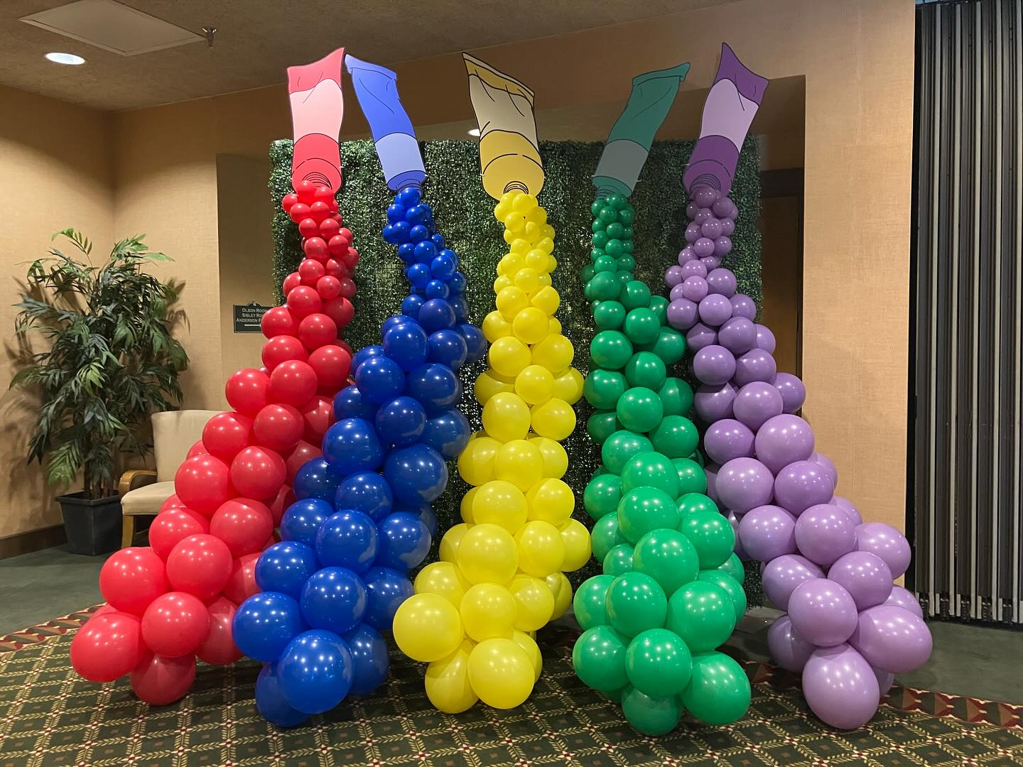 Colorful balloon sculptures resembling crayons in red, blue, green, yellow, and purple are artfully arranged indoors, set against a backdrop of leafy plants and beige walls, creating a vibrant and festive atmosphere.