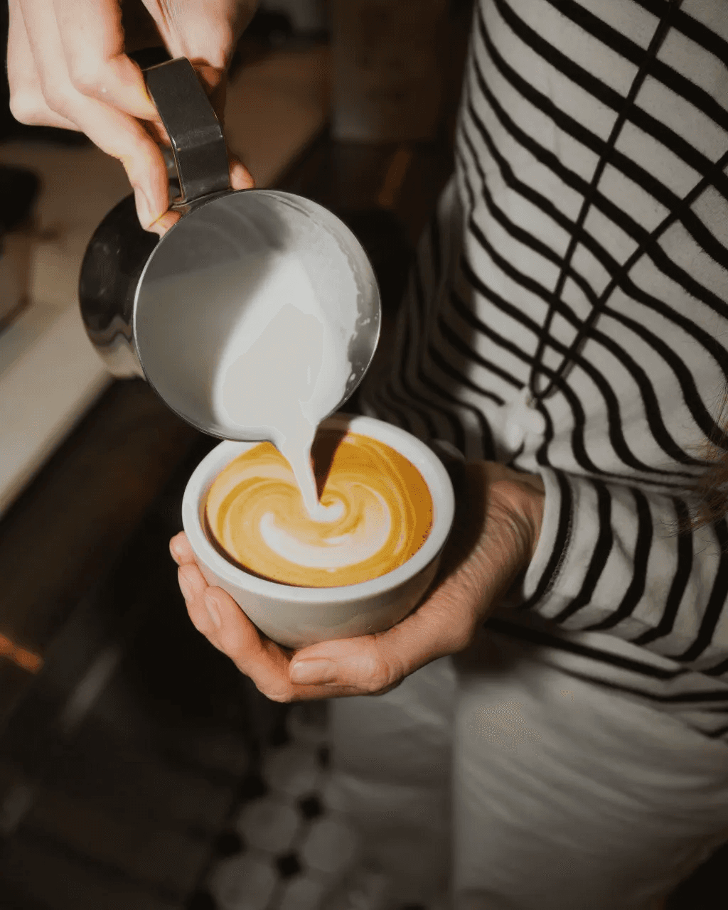 A latte with decorative foam art in a burgundy cup on a white saucer, placed on a light-colored table with a decorative plate nearby and greenery in the background