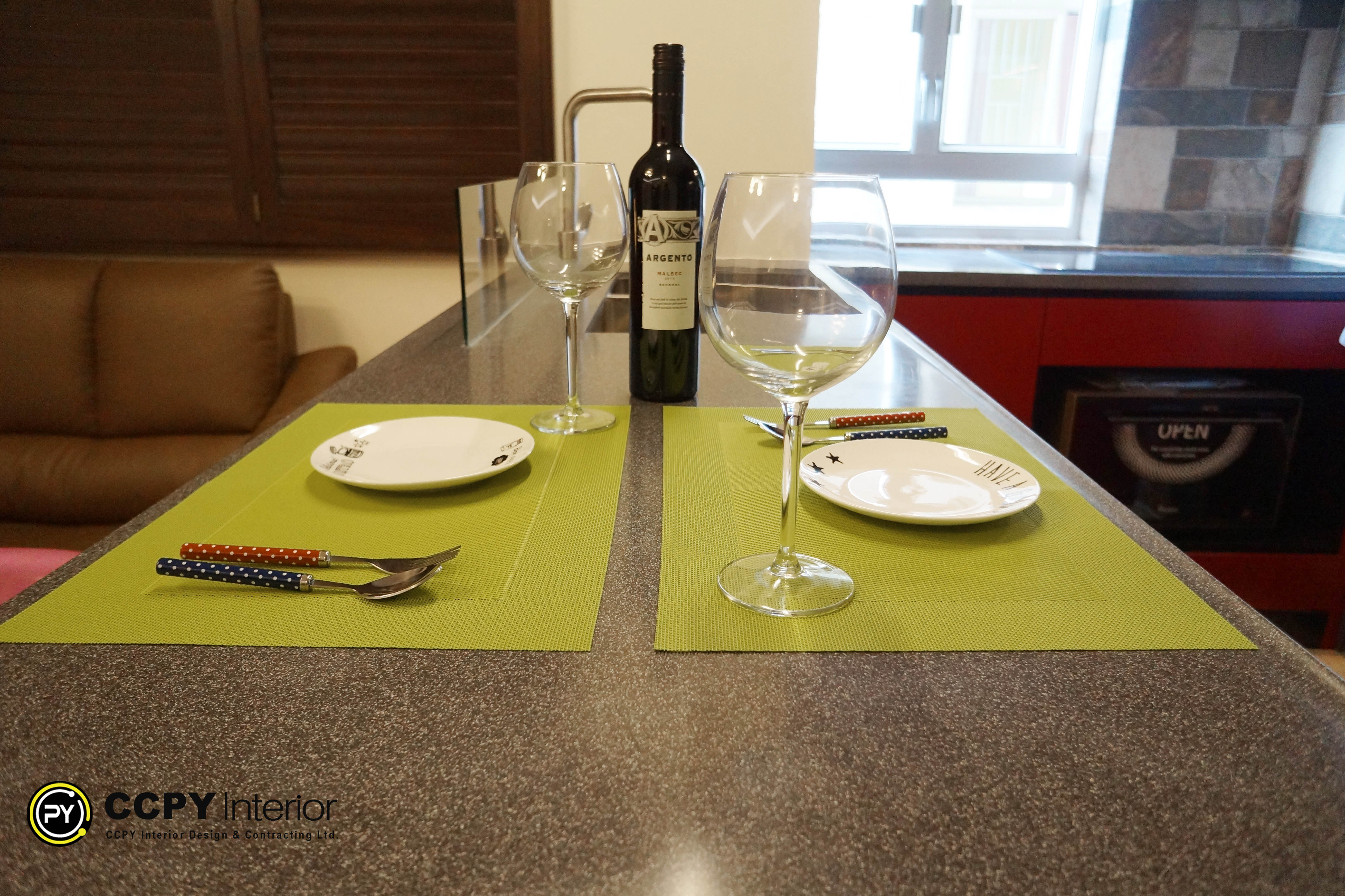 he highlighted region shows a meticulously arranged kitchen island counter that serves as a dining space.Design DetailsColor Palette: Features sharp lime-green placemats contrasted against a textured gray quartz countertop.Table Setting: Arranged for two guests with elegant crystal wine glasses, white ceramic plates, and structured silverware.Background Elements: Includes built-in dark red kitchen cabinetry, an integrated microwave oven, and a sleek modern gooseneck faucet.