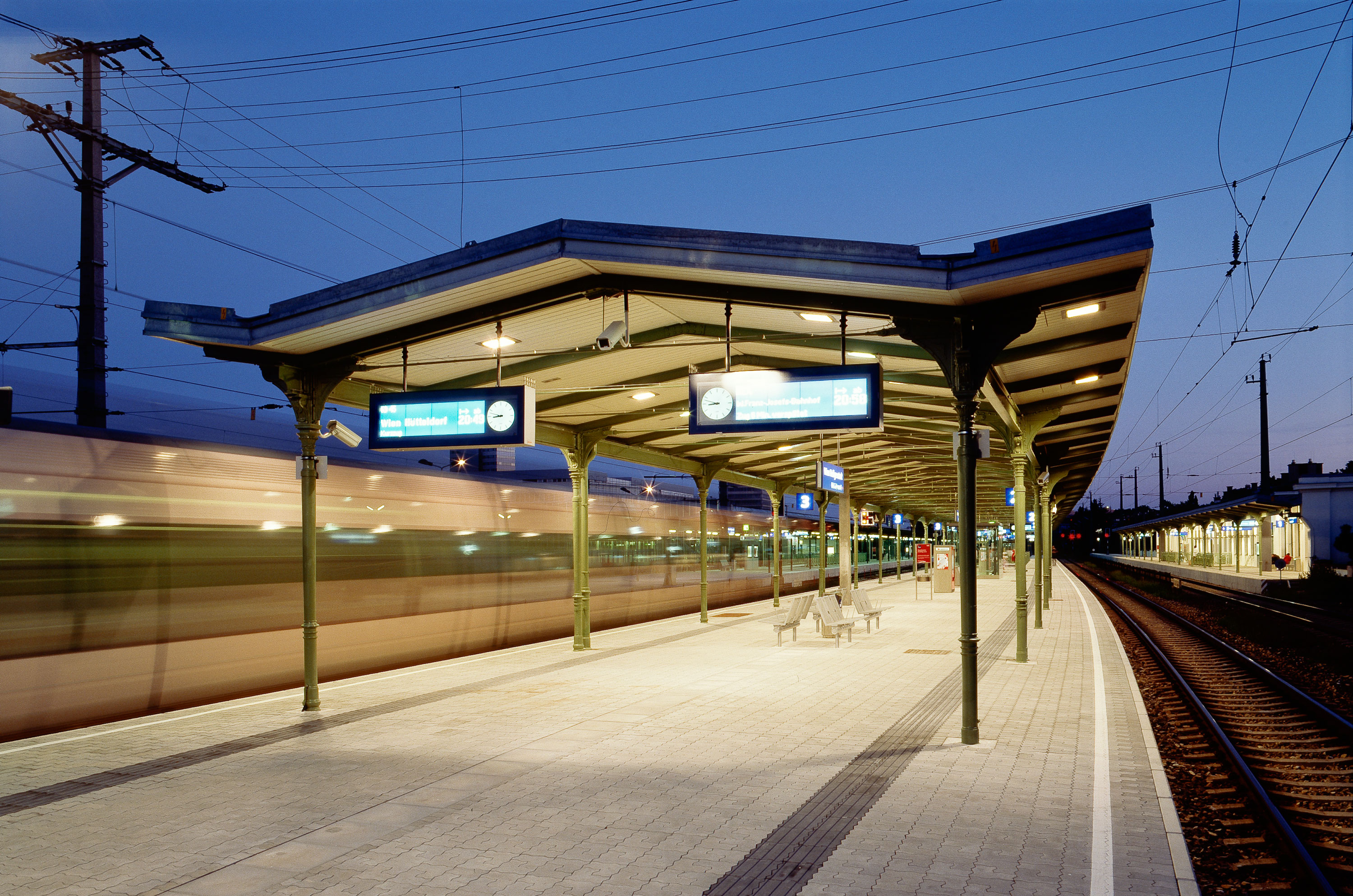Bahnsteig am Bahnhof Heiligenstadt bei Dämmerung mit Bahnsteig und historischer Überdachung, zwei beleuchteten Anzeigetafeln und vorbeifahrender Zug links.