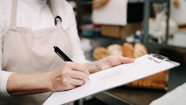 Pessoa de avental bege escrevendo à caneta em papel na prancheta, em ambiente de padaria.