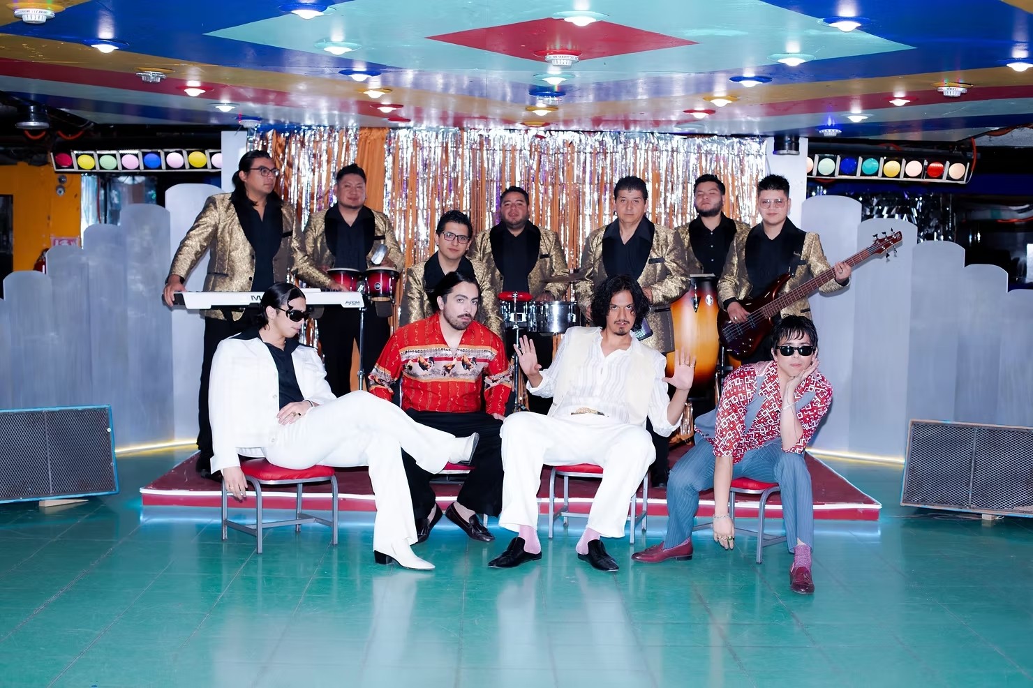 A lively music band poses on a colorful stage with shiny silver streamers. Members wear gold jackets, red and white outfits, exuding a retro vibe.