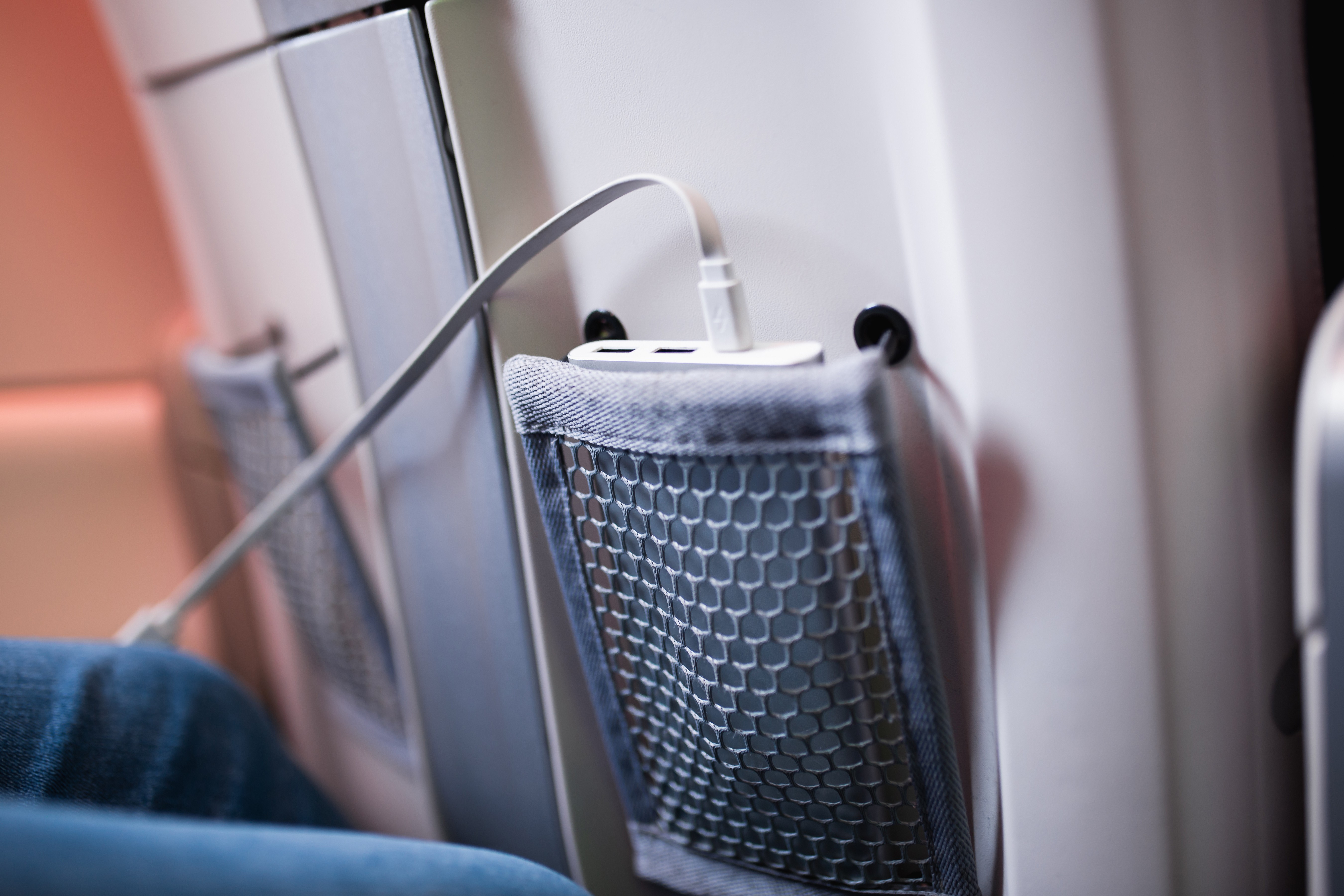 Une batterie externe blanche est rangée dans la pochette en filet du siège d'un avion, avec un câble USB branché pour charger un appareil électronique. On voit en partie l'intérieur de la cabine et un passager assis à proximité.