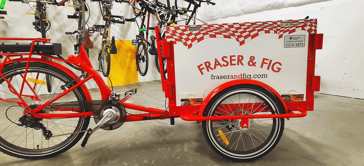 a red-and-white bicycle with fridge box attachment and vinyl wrap on top, reading Fig & Fraser on the sides and front