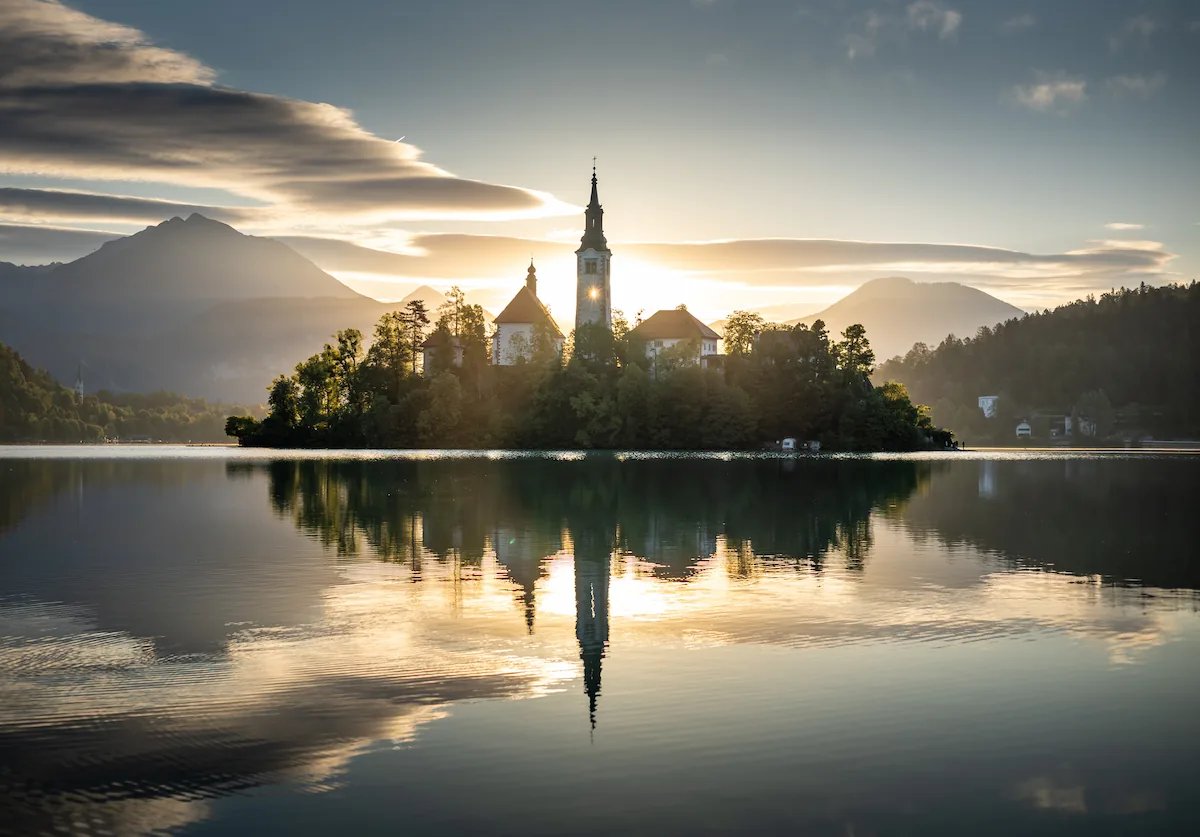 Lake Bled island church at sunrise, surrounded by still water reflecting a bright, set against the Julian Alps in Slovenia.