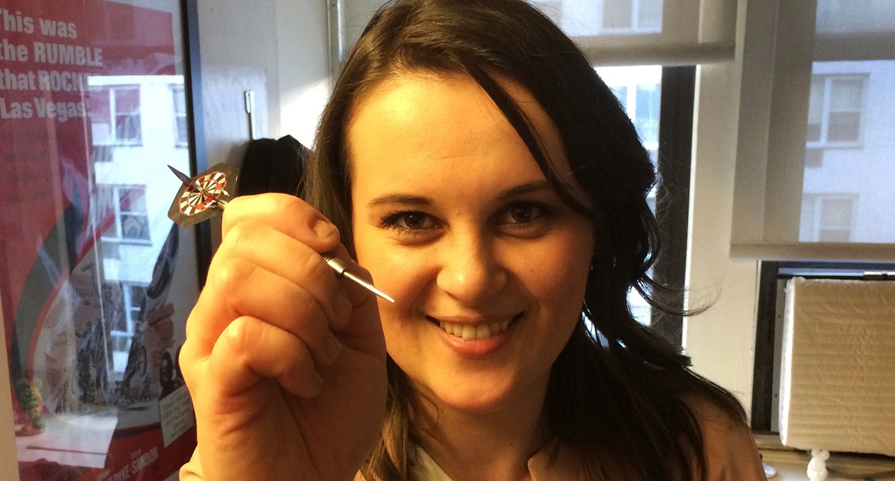 A photo of a woman holding a darts arrow in her hand.