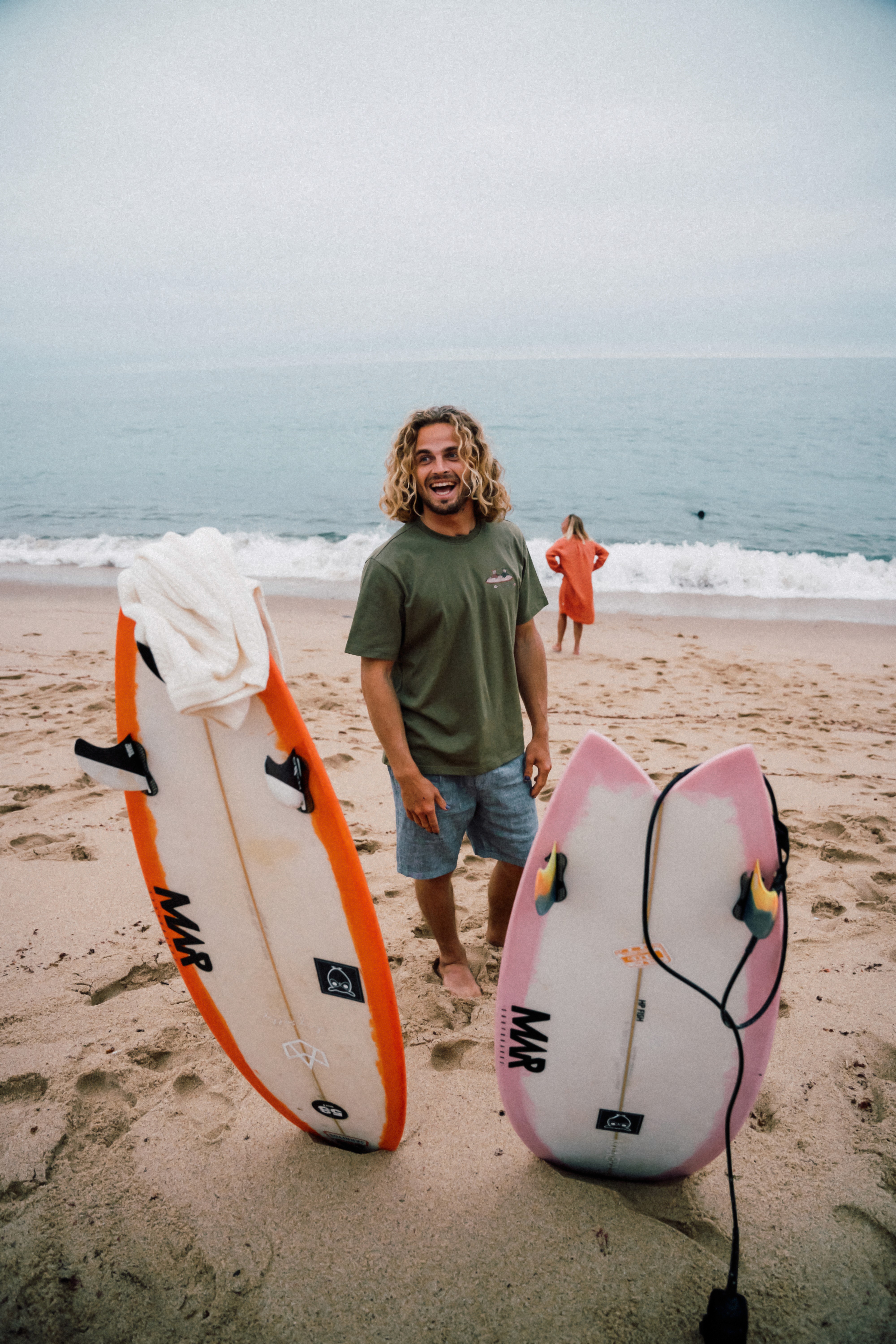 Un garçon habillé d'un t-shirt knowledge cotton apparel pose devant la mer à côté de planche de surf