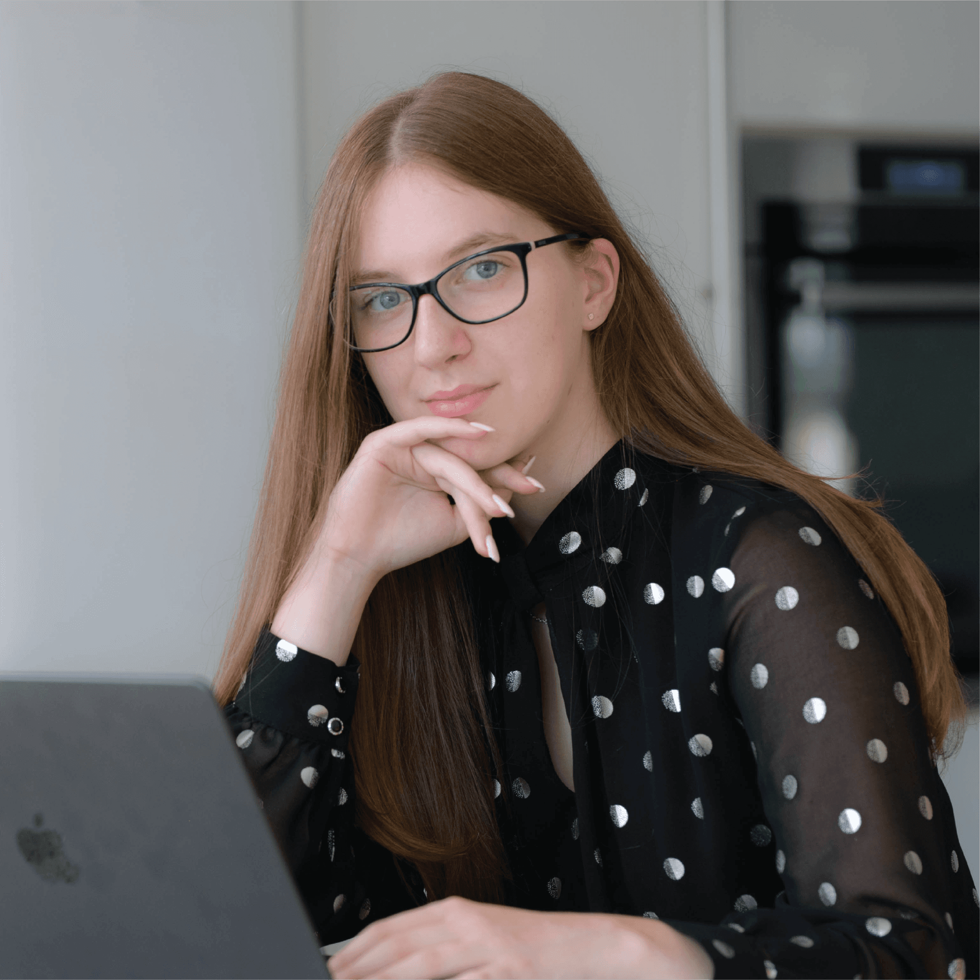A young person with curly hair smiles at the camera while sitting in front of a laptop against a dark background.