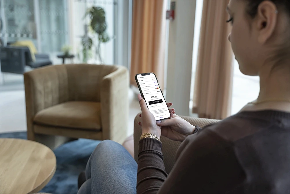 A young person with curly hair smiles at the camera while sitting in front of a laptop against a dark background.