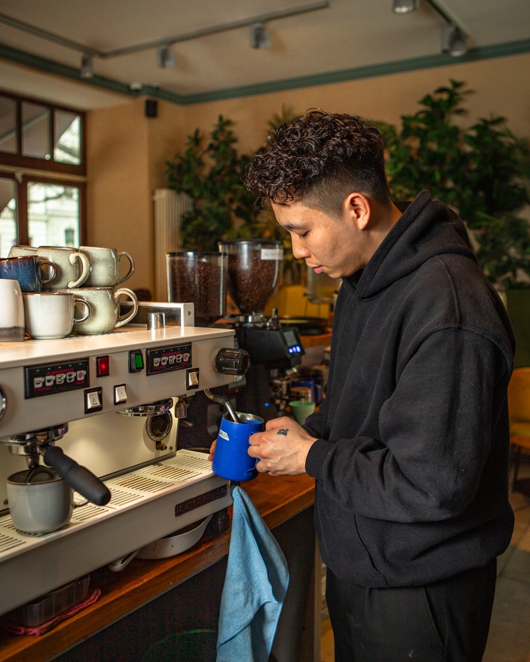 Ein Barista in einem schwarzen Kapuzenpullover dämpft konzentriert Milch in einem blauen Kännchen an einer Espressomaschine in einem Café.