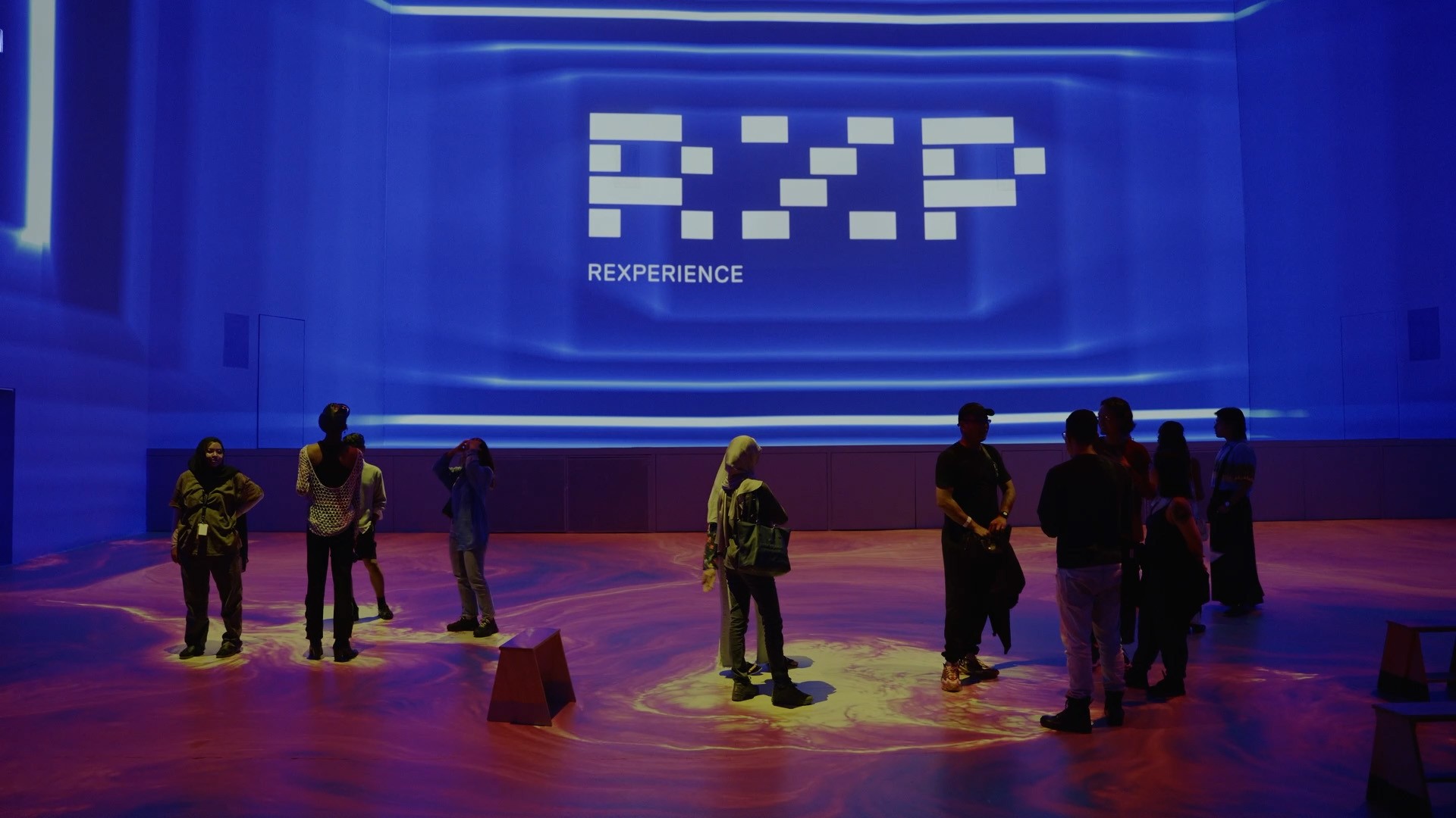 People gather in a room with a large REXPERIENCE projection