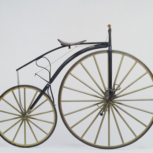 Bicicleta antigua con una gran rueda delantera, una rueda trasera más pequeña y un marco de metal negro curvado sobre un fondo blanco liso.