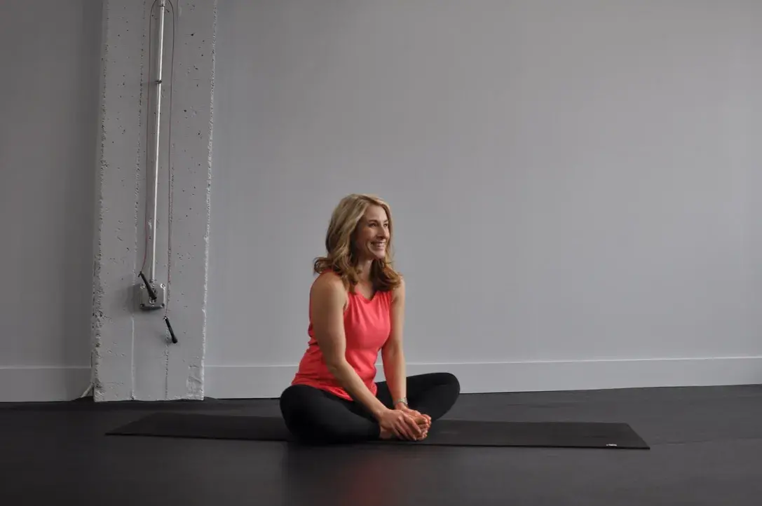 Heather Hausman in athletic wear performs a seated butterfly stretch on a black yoga mat in a minimalist, industrial-style workout space.