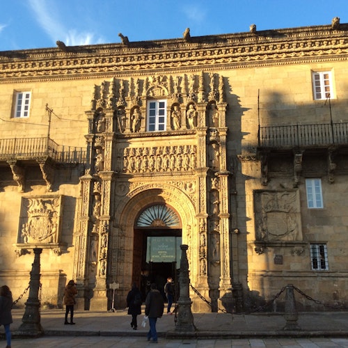 Facciata di un edificio storico ornata con statue e elaborati lavori in pietra. Persone che camminano nei dintorni sotto un cielo azzurro.