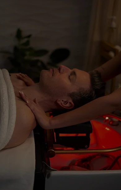 A person is lying on a massage table, covered with a towel, receiving a relaxing head and shoulder massage in a dimly lit spa environment with ambient red lighting and green plants in the background.