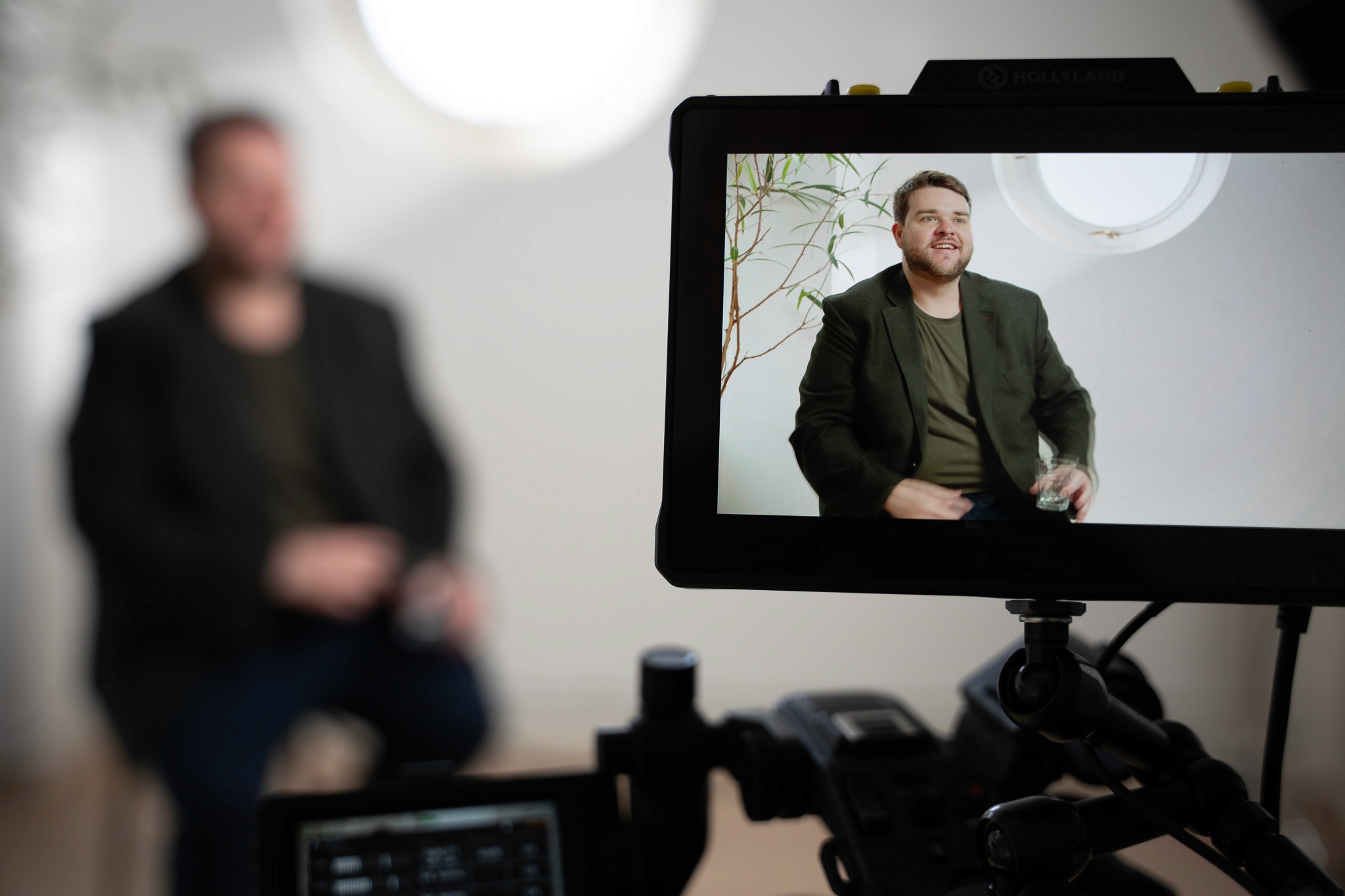 Close-up of a camera monitor showing a man in a dark blazer smiling mid-interview, with the subject blurred in the background behind the filming equipment.