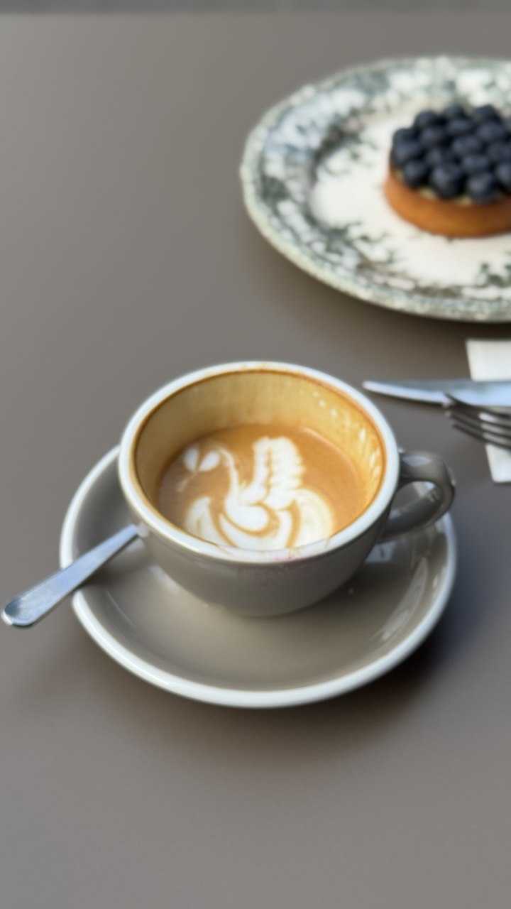 A creamy cappuccino with intricate latte art in a gray ceramic cup is placed on a saucer next to a plate topped with a blueberry tart, set on a smooth, neutral-toned table.