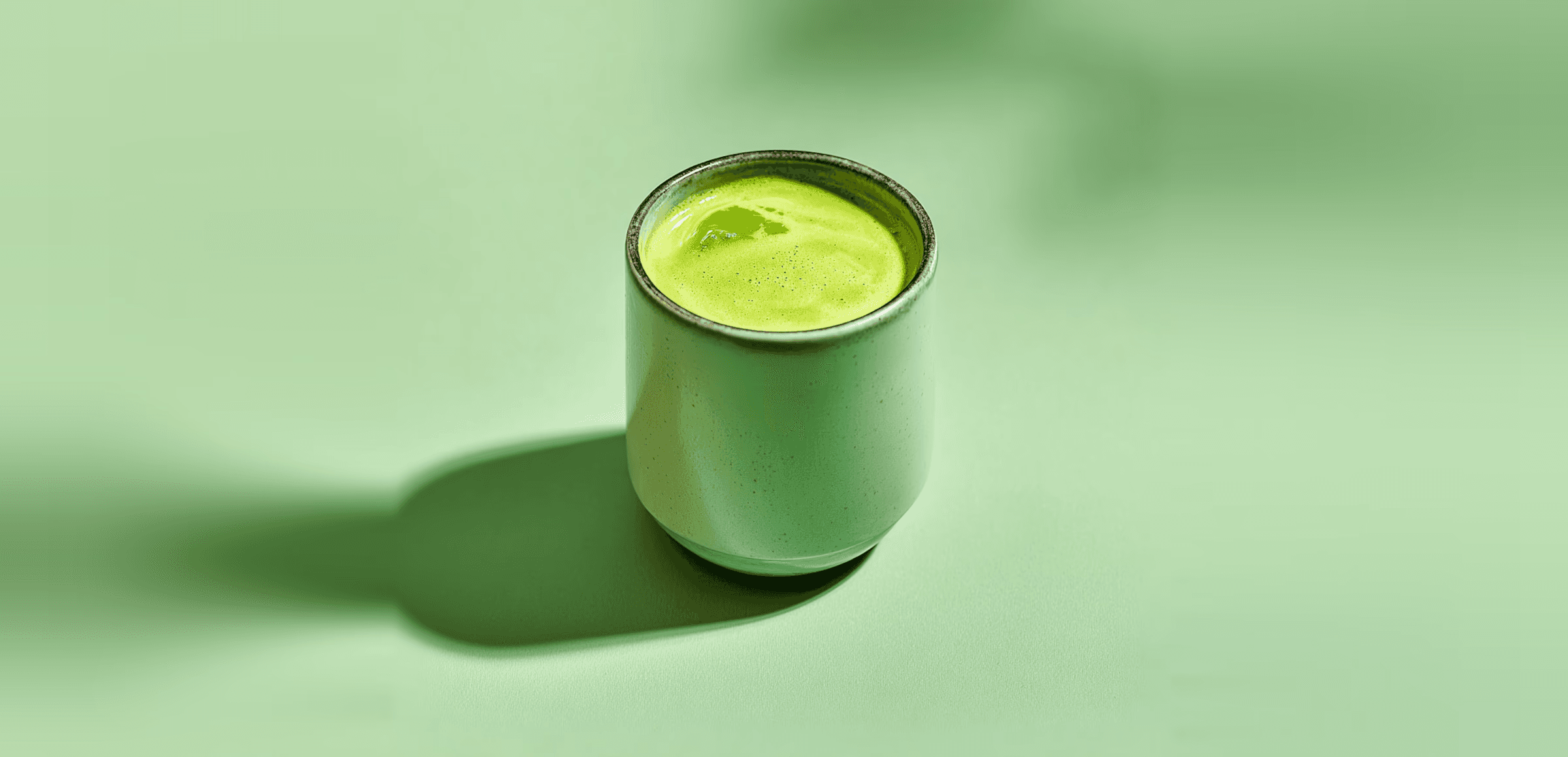 Minimalist still life of a matcha green drink in a ceramic cup on a pale green surface with long artistic shadows.