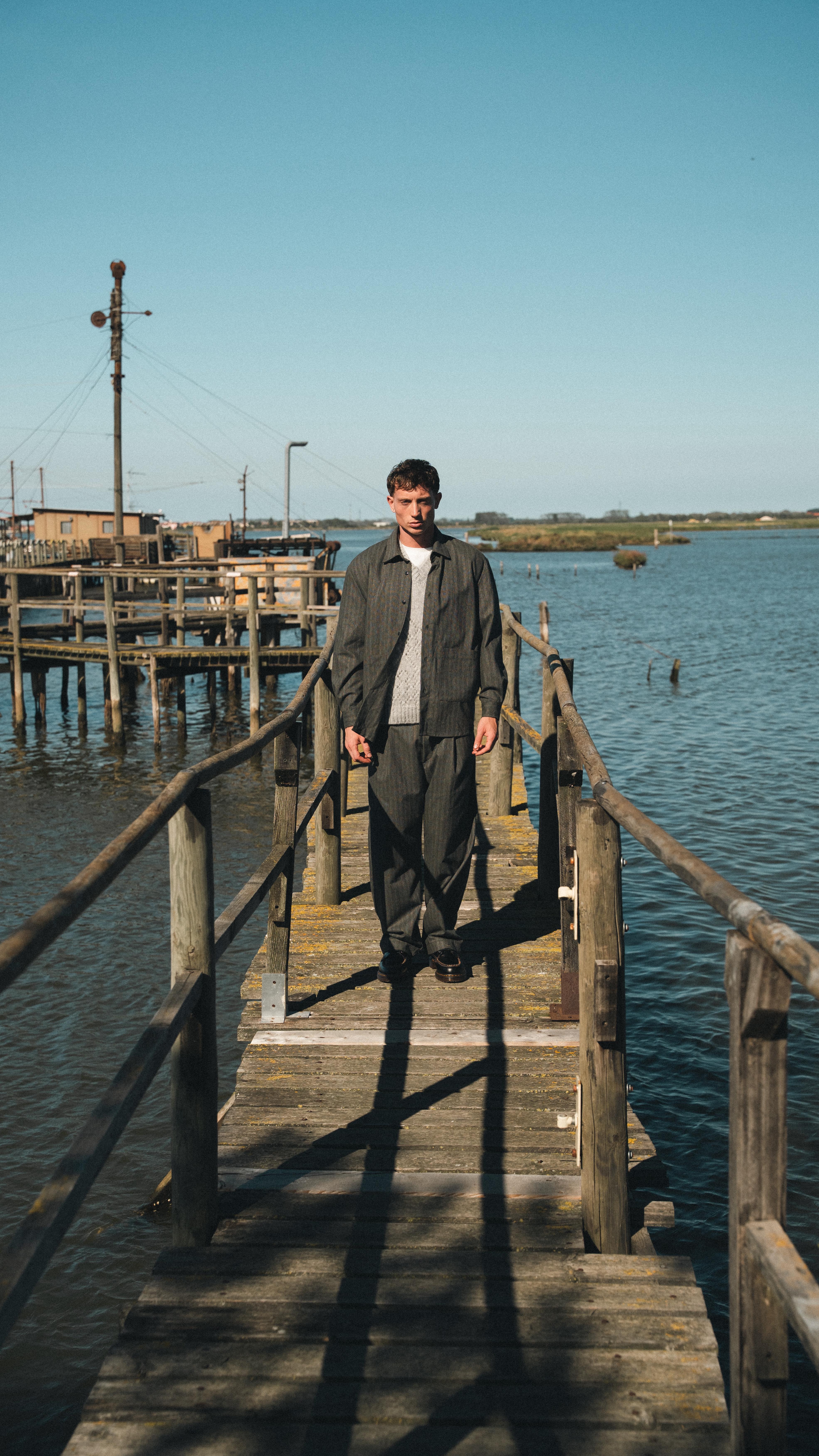 Mannequin homme sur une passerelle au dessus de l'eau, portant un ensemble pantalon et sur-chemise dans le lookbook FW25 de Wooden Store