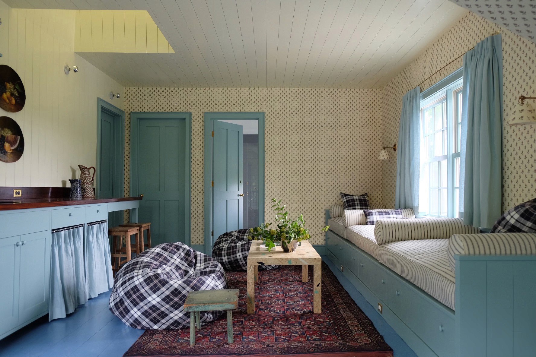 Guest cottage living room with built-in sofa, plaid bean bags, and patterned wallpaper.