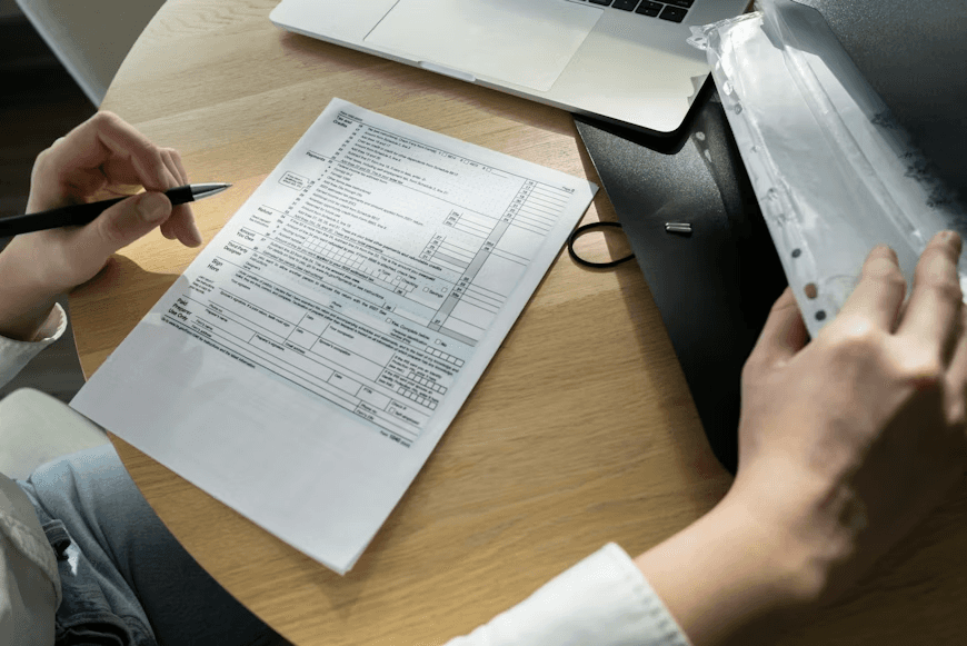 A close-up of someone at a desk reviewing a printed form, holding a pen while organizing papers in a clear plastic sleeve next to a laptop.