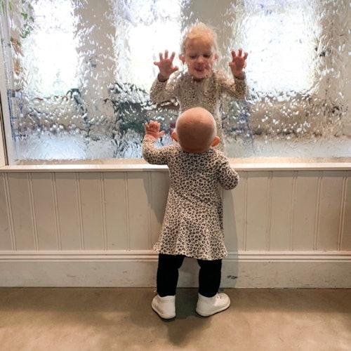 Two small children in patterned outfits play at a window with textured glass. One child stands, while the other looks inside.