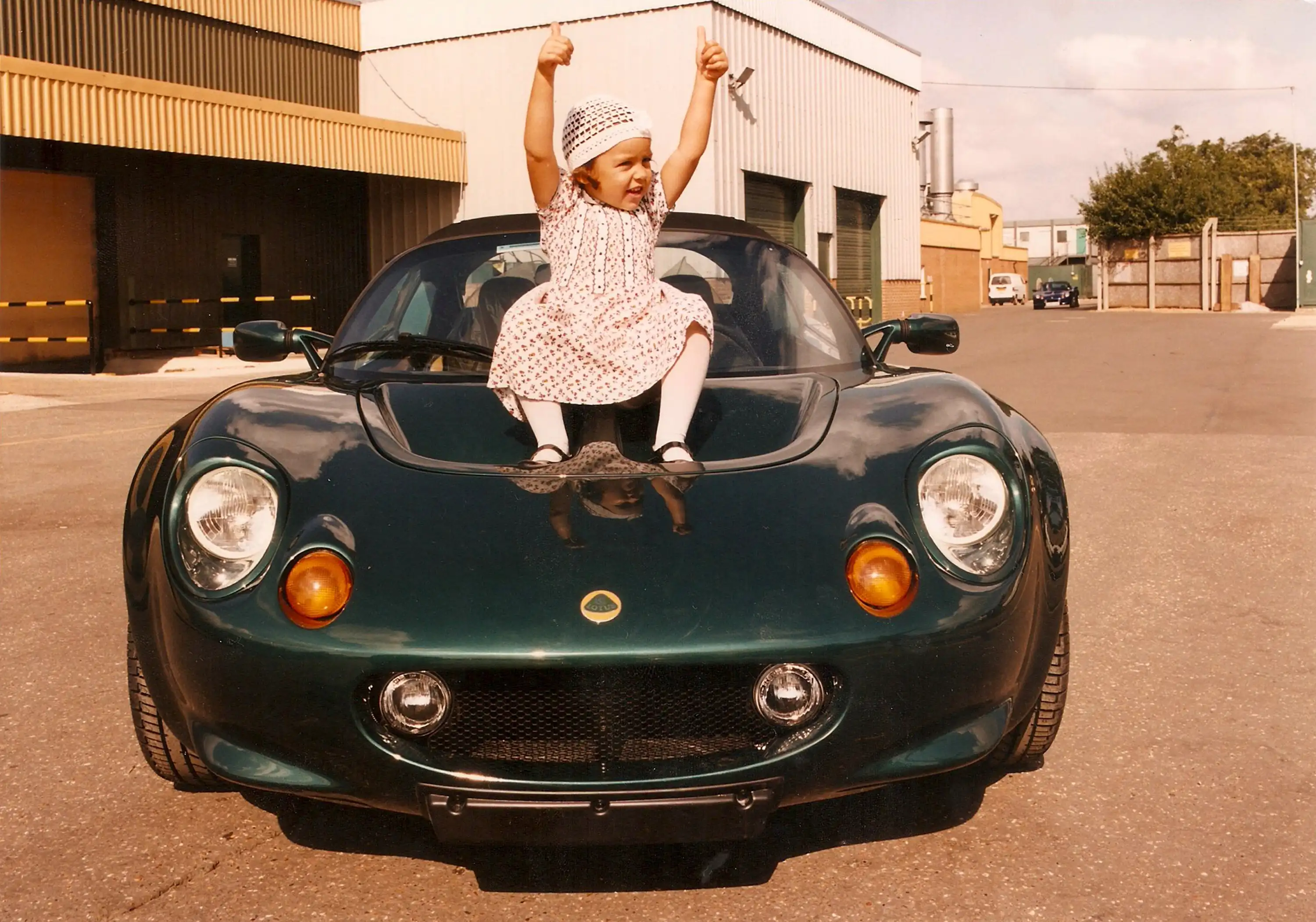 A young Elisa Artioli in a patterned dress and hat sits on the hood of a dark green Lotus Elise sports car, smiling and giving two thumbs up in an industrial area.