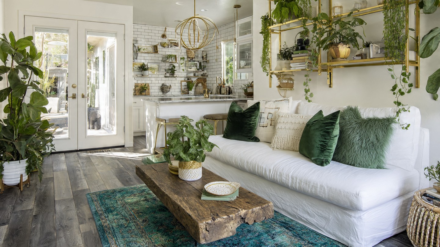 Bright living room with white sofa adorned by green cushions, surrounded by lush plants. Sunlight streams through French doors, illuminating the cozy, modern space.