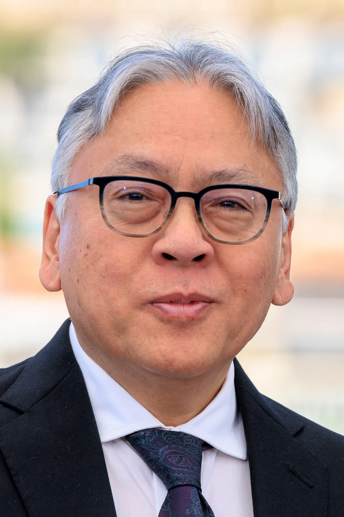 A middle-aged Asian wearing spectacles and a dark suit with white shirt, poses in an outdoor setting.