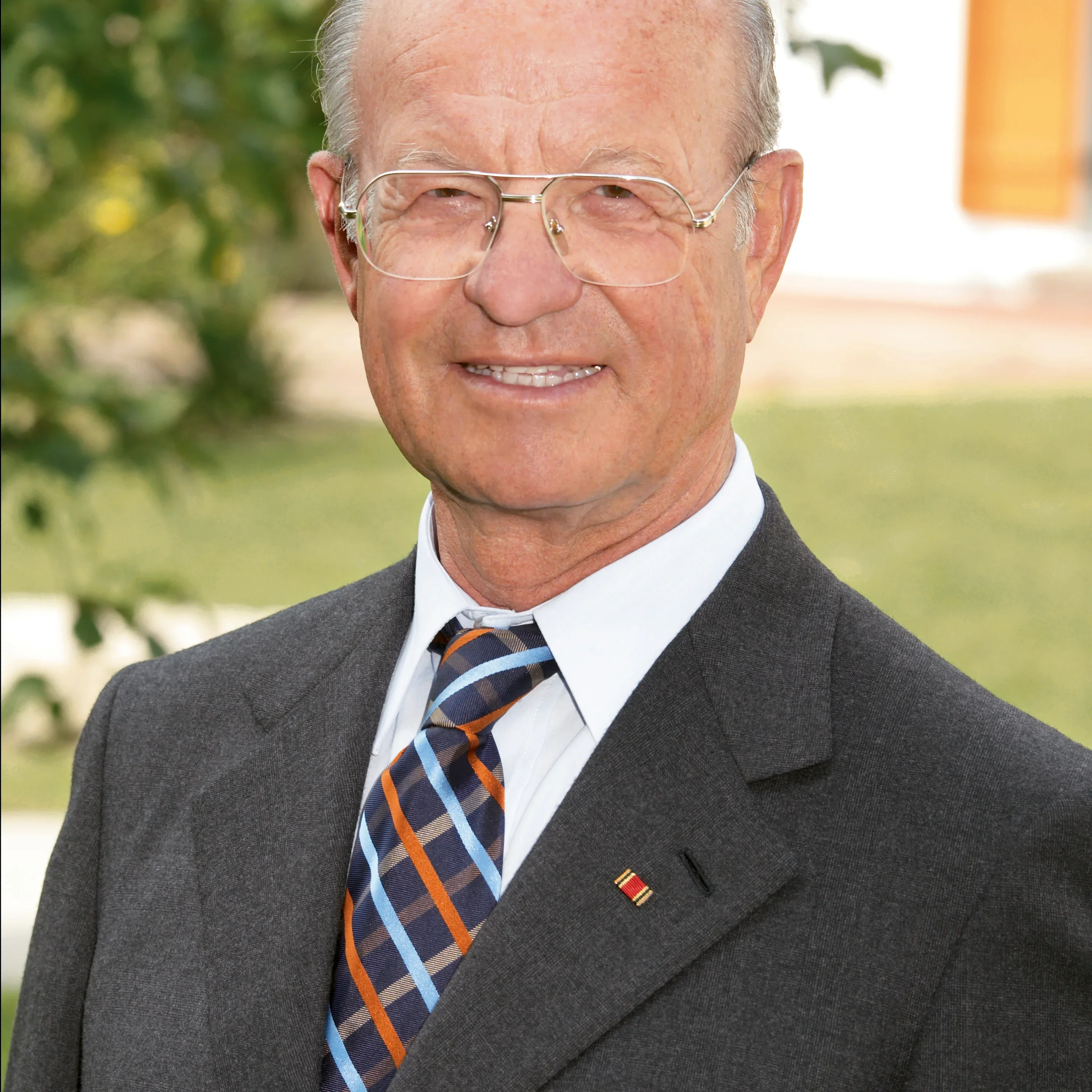 Ein älterer Herr mit Brille und schütterem grauem Haar tragend einen dunklen Anzug mit einem gemusterten dunkelblauen, orangefarbenen und hellblauen gestreiften Schlips. Im revers des Anzugs ist eine kleine rote, gelbe und schwarze Auszeichnung angeheftet. Er lächelt leicht in Richtung des Betrachters vor einem unscharfen grünen Hintergrund, der möglicherweise einen Garten darstellt.