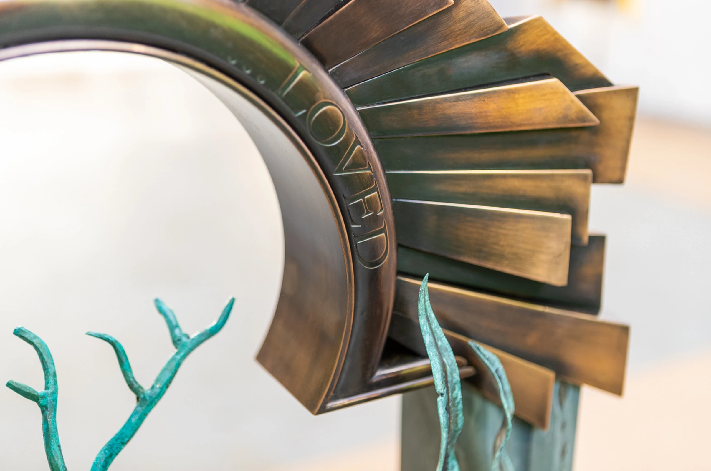 A close-up detail of a bespoke polished bronze headstone sculpture engraved with text, featuring green patinated bronze leaf elements. A tailored memorial objet d’art piece by Shaun Leane.