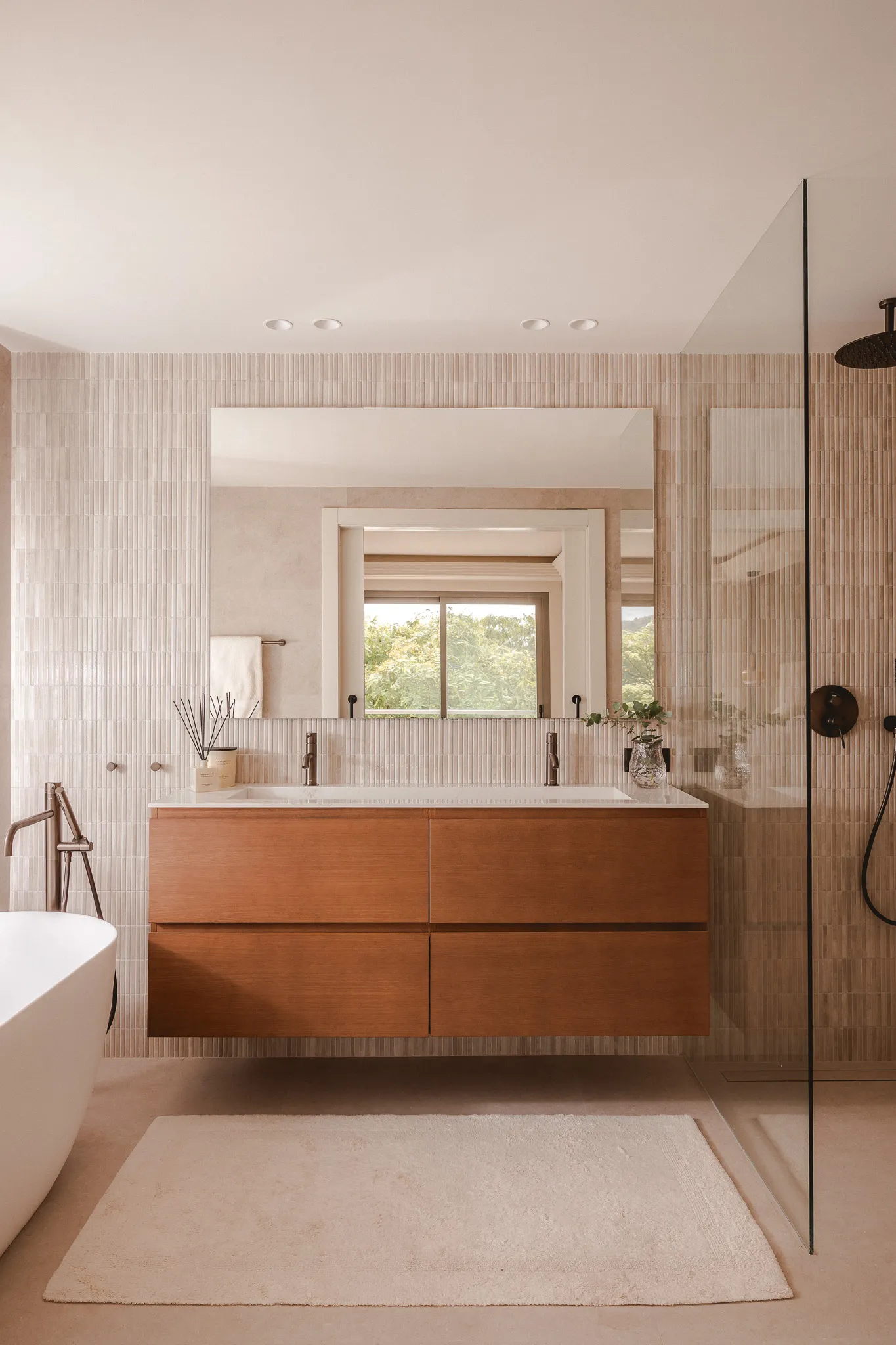 A modern bathroom featuring a floating wood vanity with a double sink, a large frameless mirror, vertical beige kit-kat tiles, a white rug, and a partial view of a freestanding bathtub and glass shower.