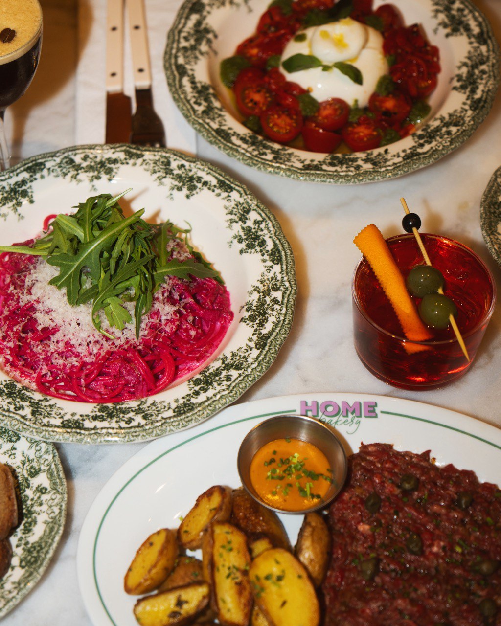 An overhead shot of a restaurant table displaying multiple dishes on decorative vintage-style plates with ornate green borders. The dishes include: beetroot pasta with fresh arugula and grated cheese (top left), roasted cherry tomatoes with burrata cheese and basil (top right), a red cocktail garnished with carrot stick and olives (center right), and beef tartare with potato wedges and sauce in a small bowl on a plate marked "HOME" 