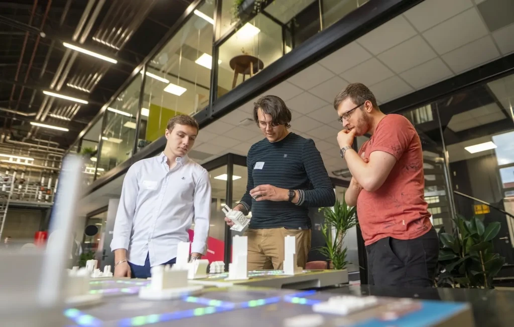 Three young men studying an illuminated architectural city scale model in a modern co-working space