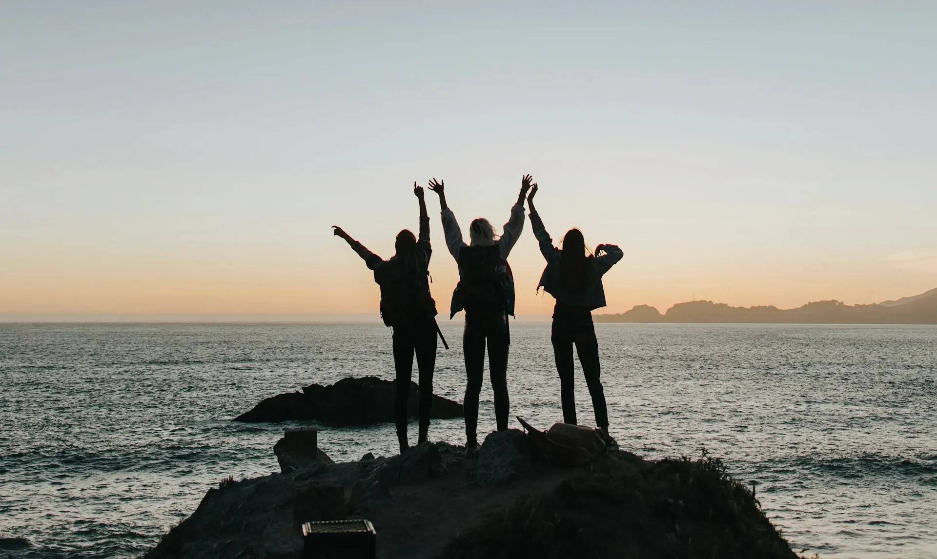 Three people standing on a rocky coastline with arms raised, silhouetted against a sunset over the ocean.