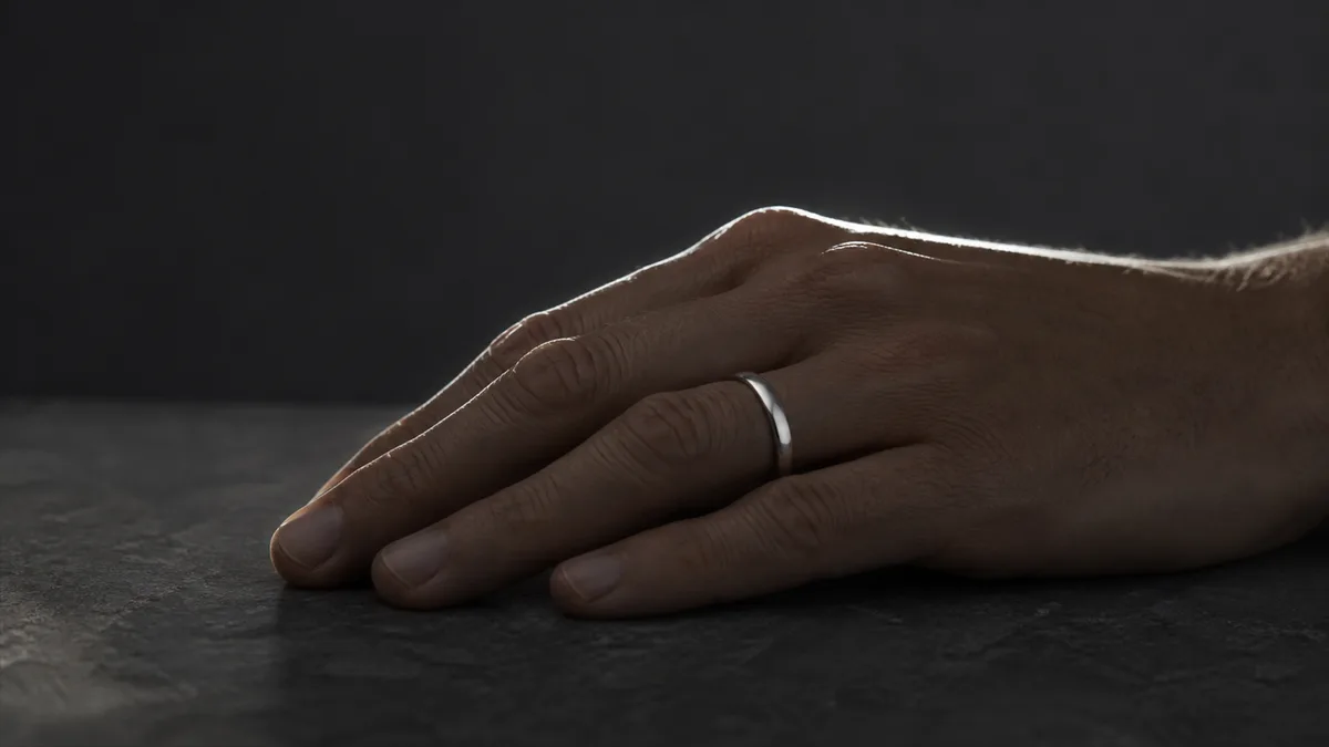 A relaxed hand wears a plain silver ring in silhouette against a charcoal background