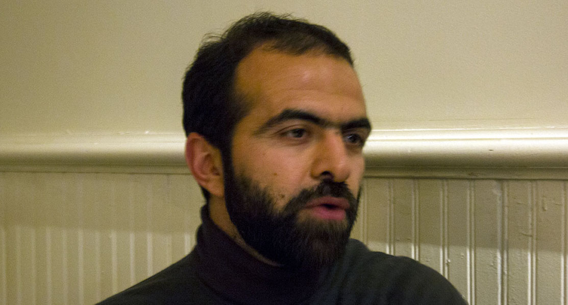 A photo of a man sitting in front of a white wall with patterns on it.