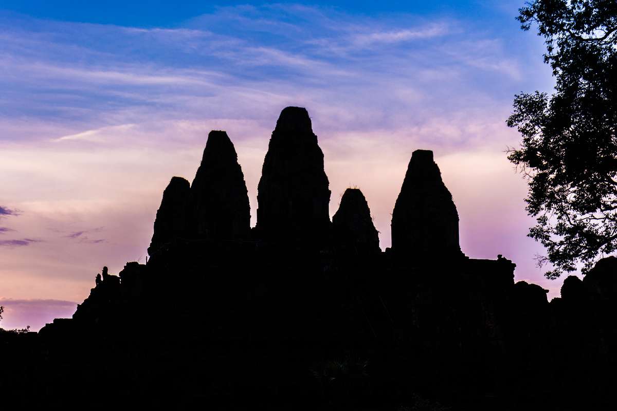 Silhouette of ancient stone temple ruins against a blue and pink sunset sky with a tree on the right.