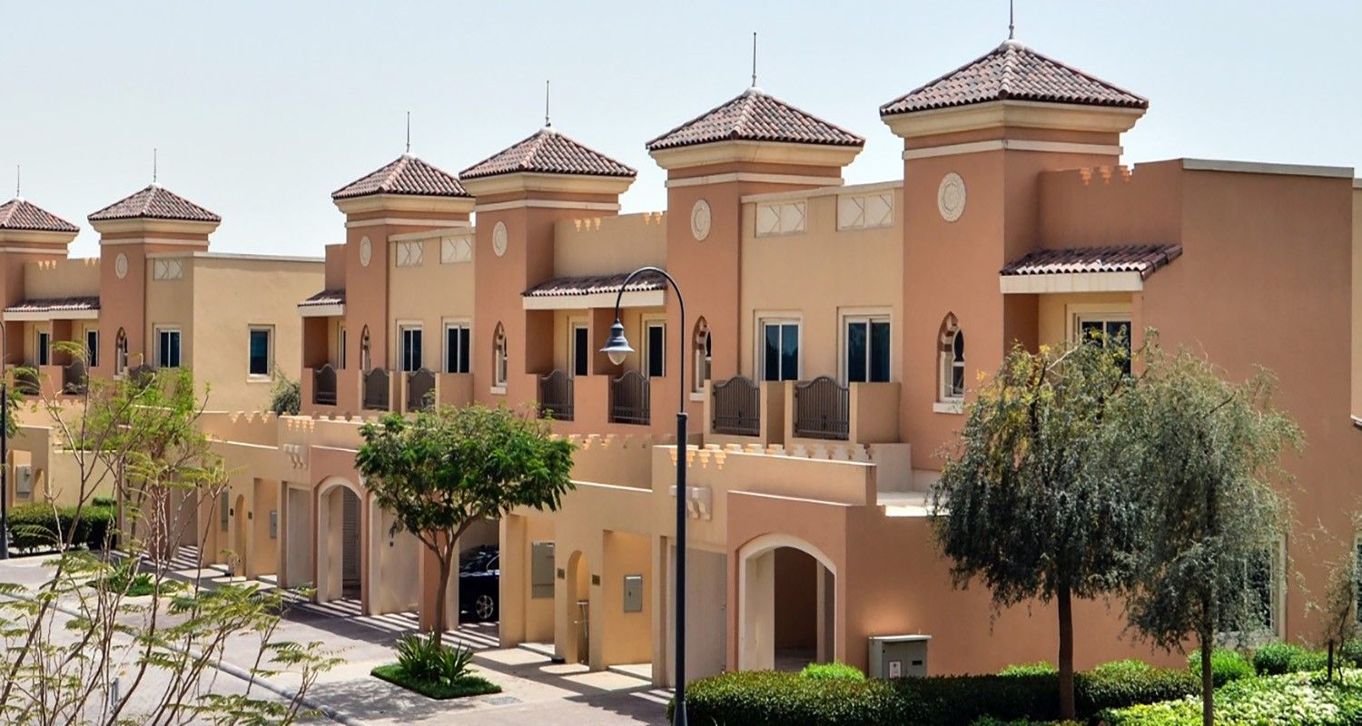 A row of townhouses at Victory Heights at Dubai Sports City, featuring beige exterior walls, balconies, and landscaped greenery. 