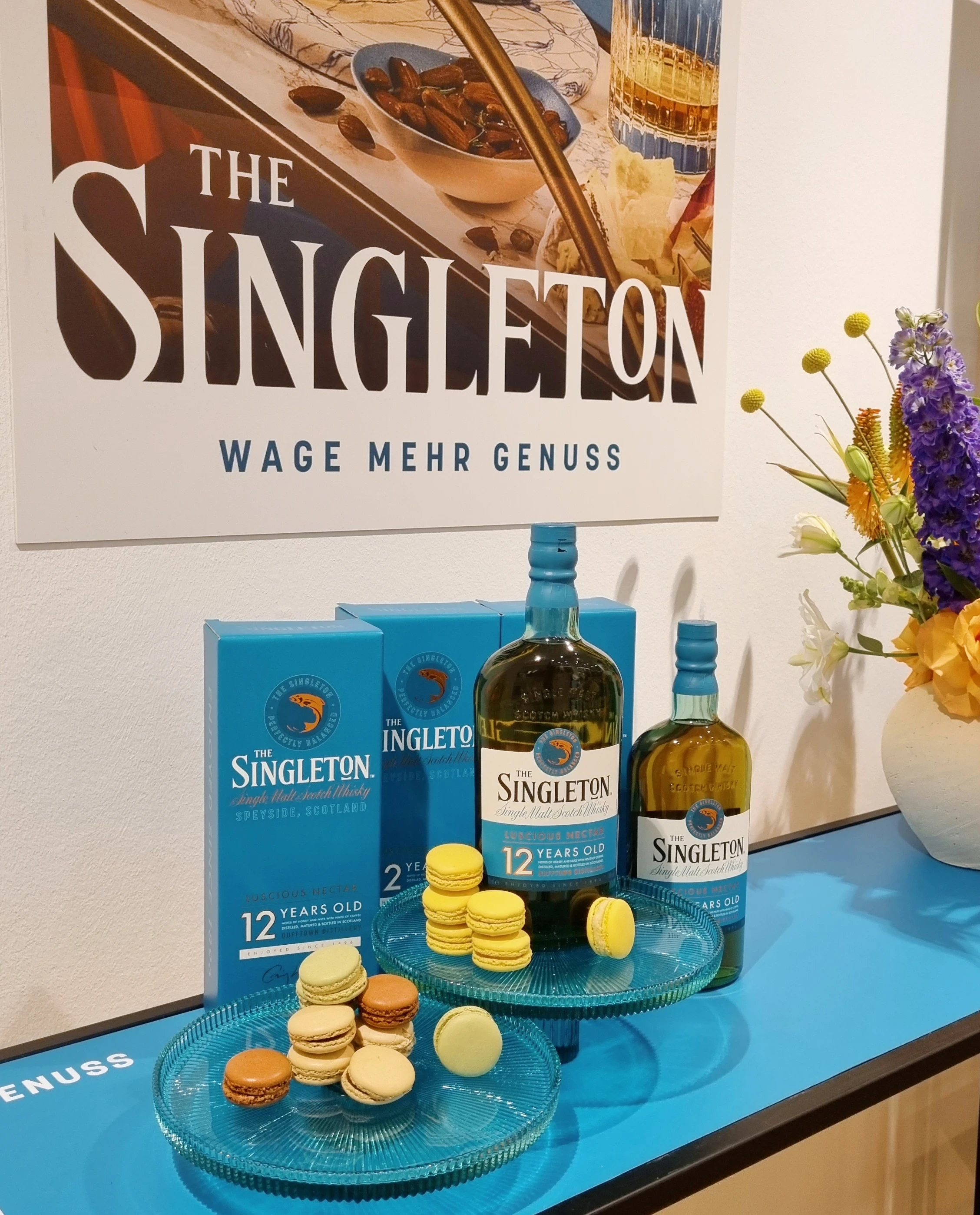 Singleton whisky bottles and packaging with macarons on glass plates in front of a campaign poster.