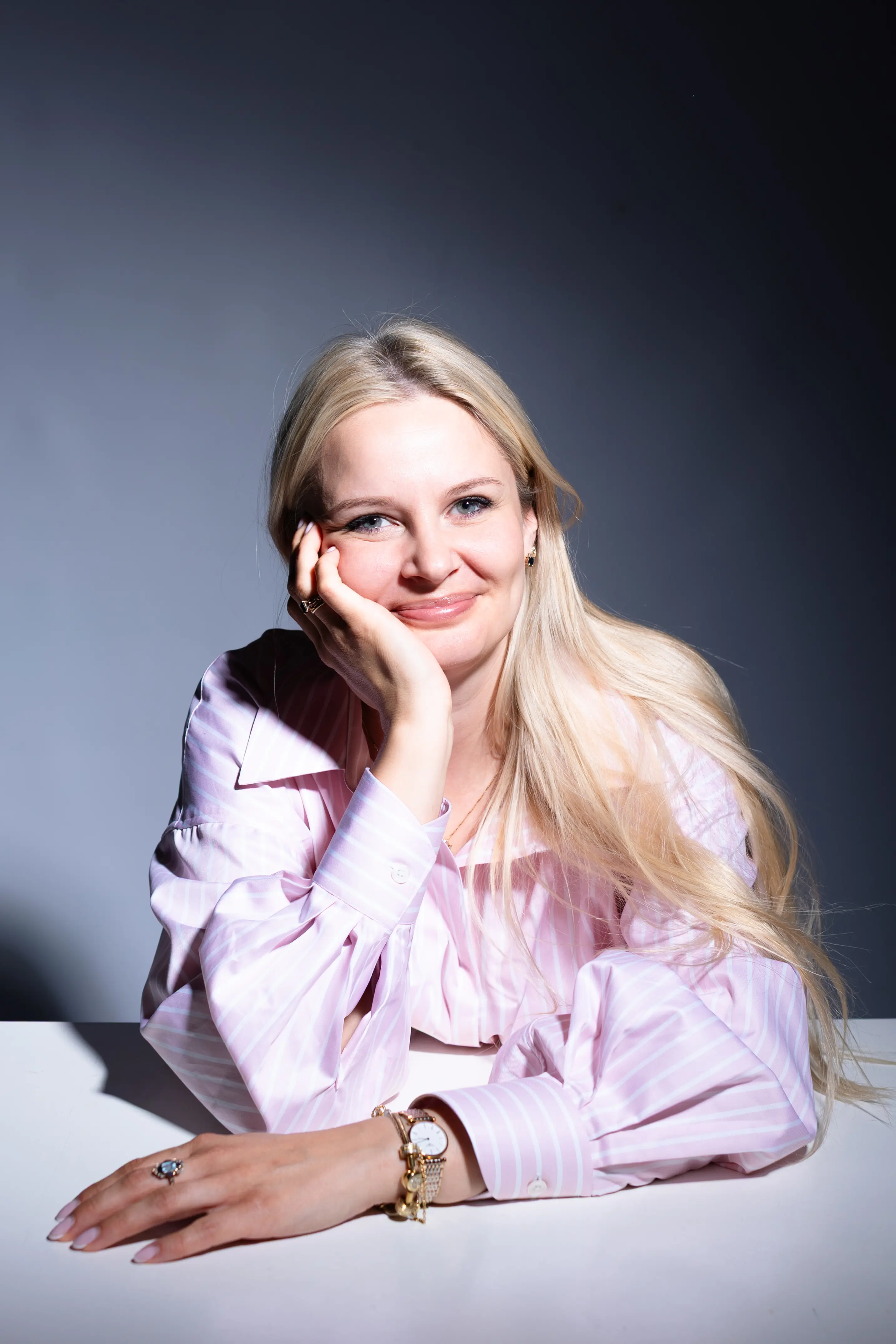 Kobieta o blond włosach w różowej koszuli, uśmiecha się i opiera o jasny stół.