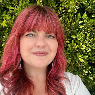 A person with vibrant red hair smiles, standing in front of lush green foliage.