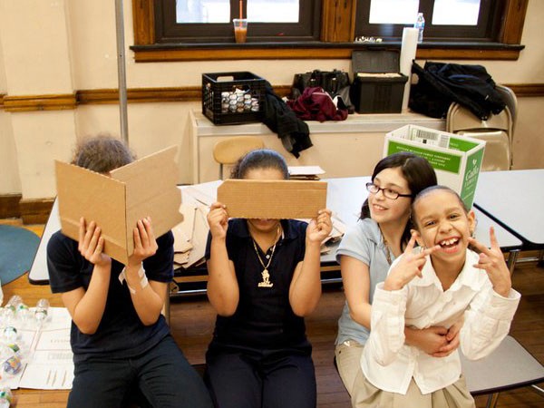 Four girls are seated on chairs in a classroom, each holding a piece of cardboard in front of their faces. The girl on the far left wears a dark blue shirt and dark pants. The girl next to her wears a black shirt and dark pants. The third girl wears a light blue shirt and glasses. The girl on the far right wears a white shirt and khaki pants. All four girls are smiling and making peace signs with their hands. The classroom has wooden floors and tables with chairs. A window with wooden trim is visible in the background, along with a black plastic crate and some other items on the windowsill.