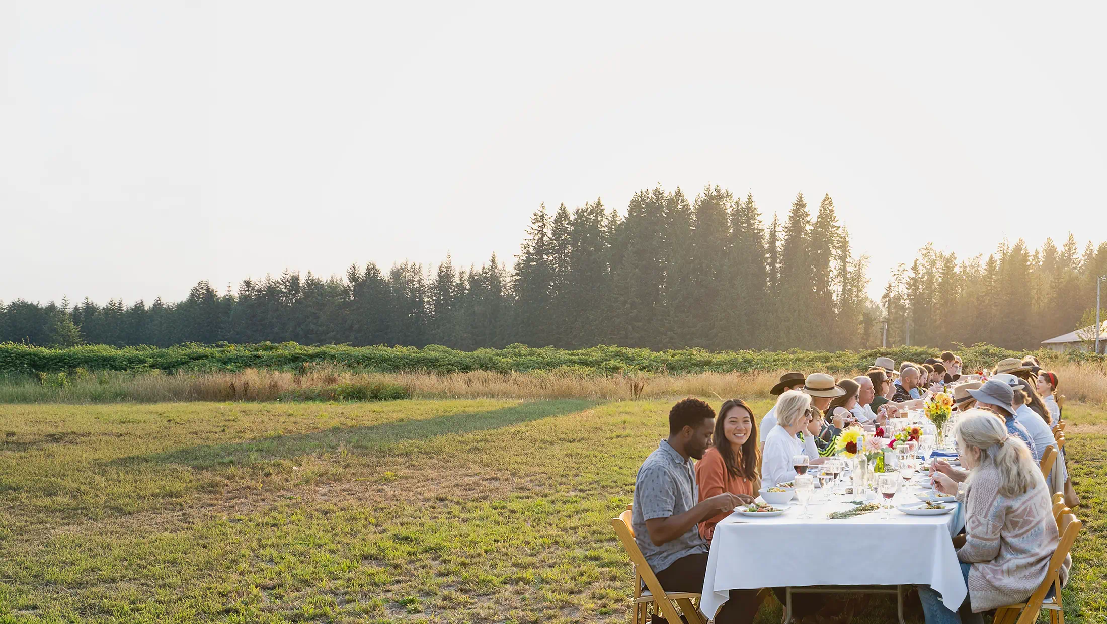 Residents and guests sharing a long outdoor meal together on farmland at Rooted Northwest.