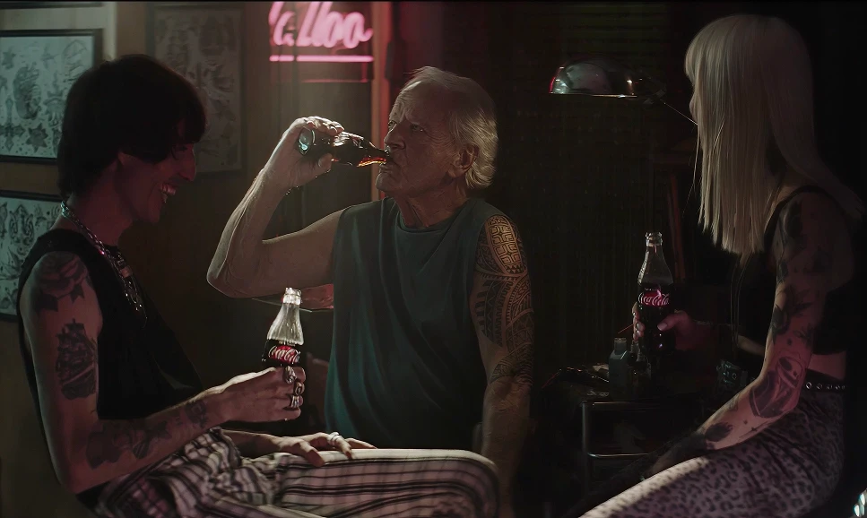 Three tattooed people sharing Coca-Cola glass bottles in moody neon-lit tattoo parlour, older man with sleeve tattoo drinking Coke flanked by two women, cinematic TV advertisement still