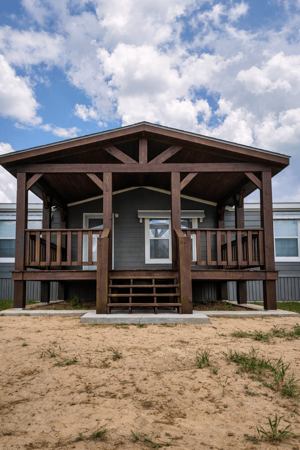 A modern, glass-walled house with warm interior lighting is perched on a rocky cliff overlooking a calm sea at sunset, under a clear blue sky.