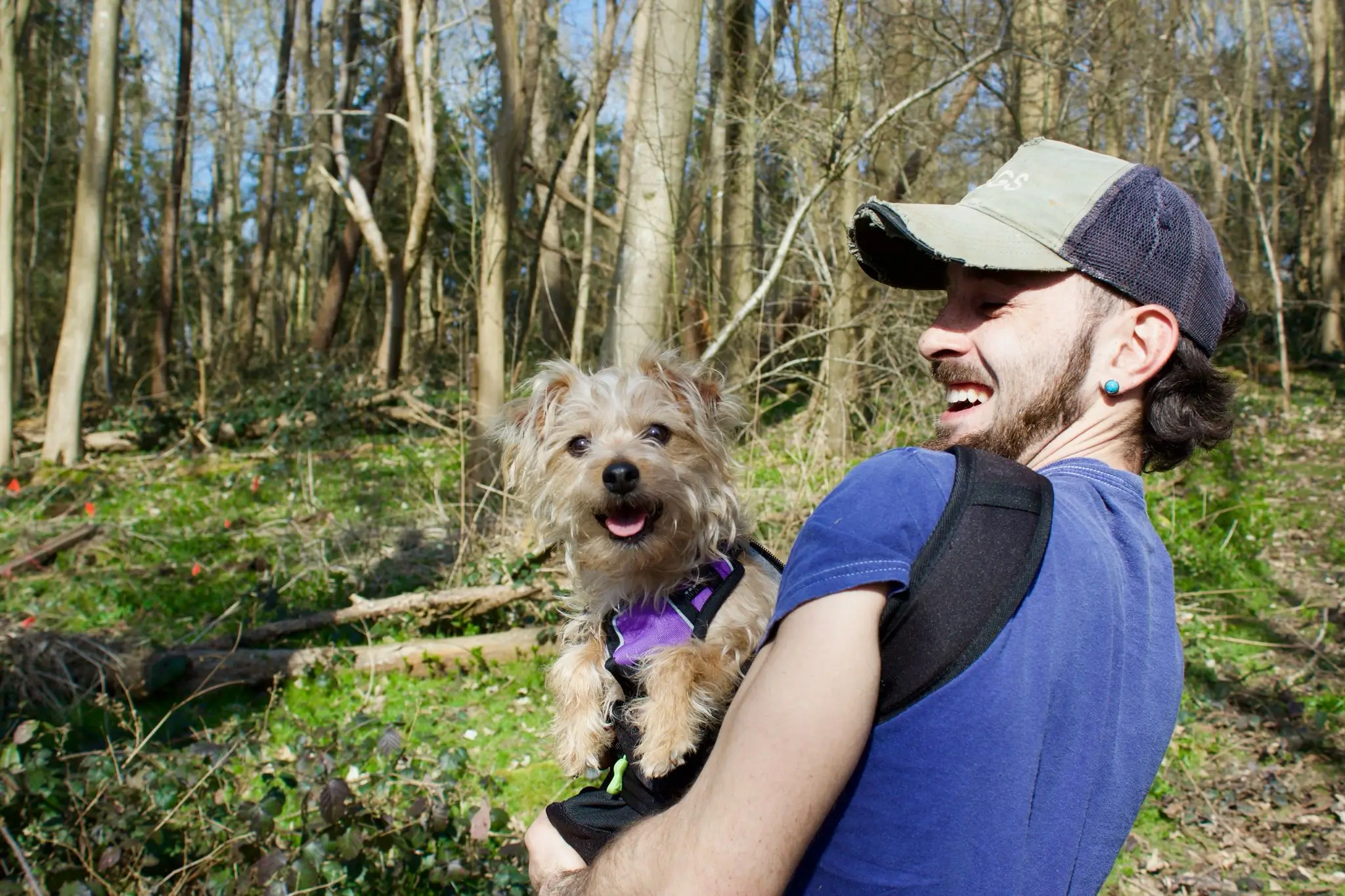 A person wearing a cap smiles while holding a small dog outdoors in a wooded area.