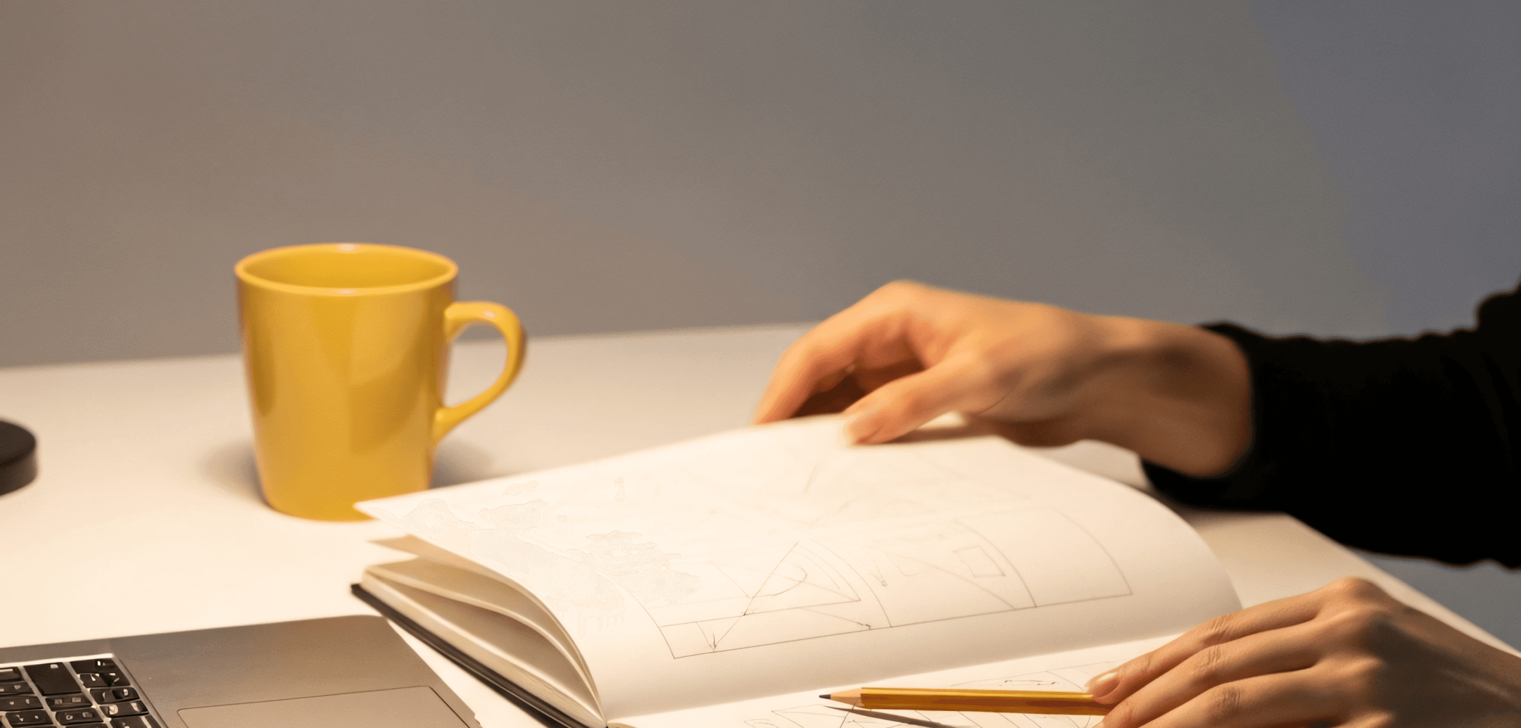 Person sketching geometric shapes in a notebook at a desk with a laptop and coffee mug